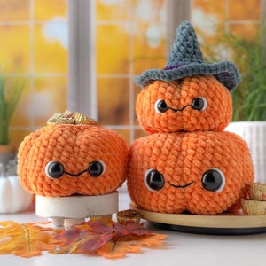 Op de afbeelding: Drie oranje pompoenen van pluche met zwarte ogen en lachende gezichten. Eén pompoen heeft een grijze heksenhoed. De pompoenen zijn gestapeld op een wit oppervlak met herfstbladeren en kleine traktaties.
