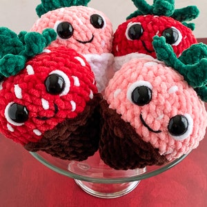 Peut inclure: Quatre jouets en peluche en forme de fraise au crochet avec un fond enrob&eacute; de chocolat. Les fraises sont roses, rouges et blanches avec des yeux noirs et une bouche souriante.