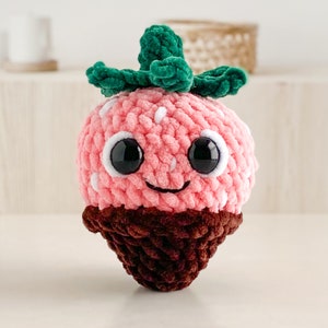 Peut inclure: Un jouet en peluche en forme de fraise crochet&eacute; avec un fond enrob&eacute; de chocolat. La fraise a une feuille verte sur le dessus et un visage souriant avec des yeux noirs.