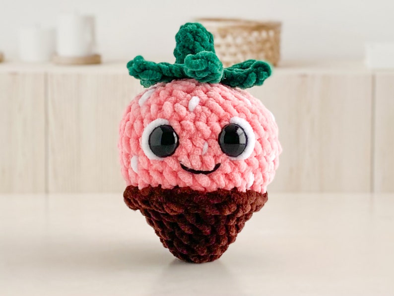 Peut inclure: Un jouet en peluche en forme de fraise crochet&eacute; avec un fond enrob&eacute; de chocolat. La fraise a une feuille verte sur le dessus et un visage souriant avec des yeux noirs.