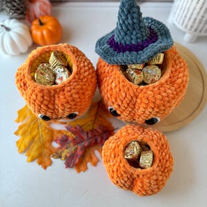 Pu&ograve; includere: Tre contenitori a forma di zucca all'uncinetto arancione con occhi mobili, pieni di caramelle incartate. Una zucca ha un cappello da strega grigio e viola. Foglie autunnali e piccole zucche sullo sfondo.