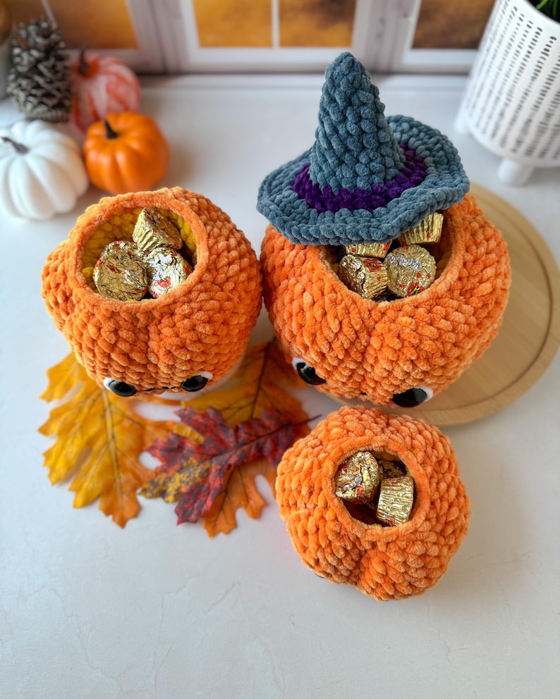 Pu&ograve; includere: Tre contenitori a forma di zucca all'uncinetto arancione con occhi mobili, pieni di caramelle incartate. Una zucca ha un cappello da strega grigio e viola. Foglie autunnali e piccole zucche sullo sfondo.