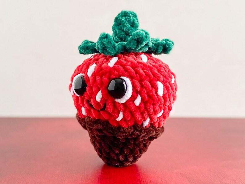 Peut inclure: Un jouet en peluche en forme de fraise crochet&eacute; avec un dessus vert feuillu, des pois blancs et un fond enrob&eacute; de chocolat. La fraise a un visage souriant avec des yeux noirs.