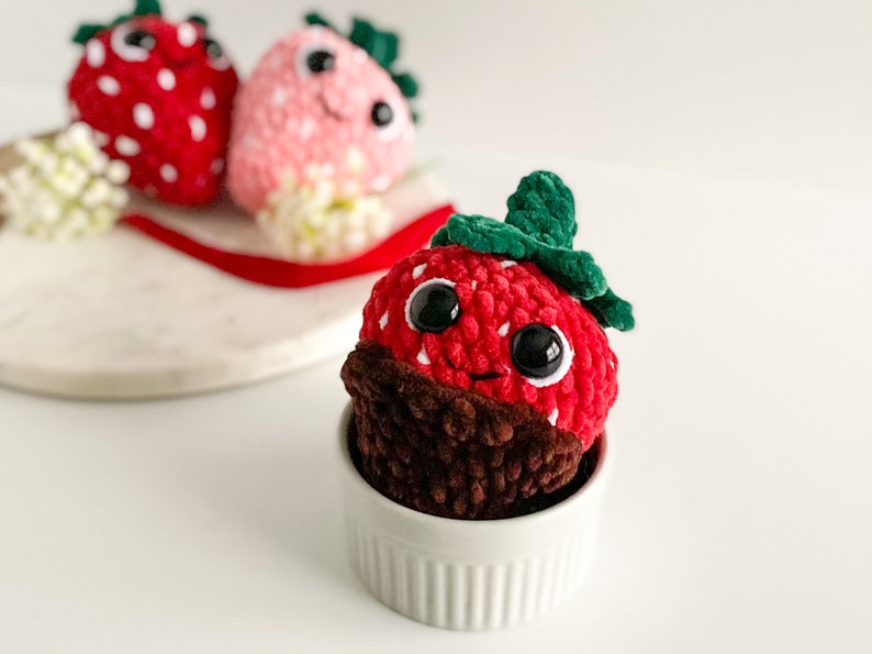 Peut inclure: Un jouet en peluche en forme de fraise crochet&eacute; avec un fond enrob&eacute; de chocolat, assis dans un bol en c&eacute;ramique blanc. La fraise a une feuille crochet&eacute; verte et des yeux en boutons noirs.