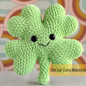 May include: A green crocheted four-leaf clover with a smiling face and black eyes. The clover is sitting on a colorful background with the text "Four Leaf Clover Modification".