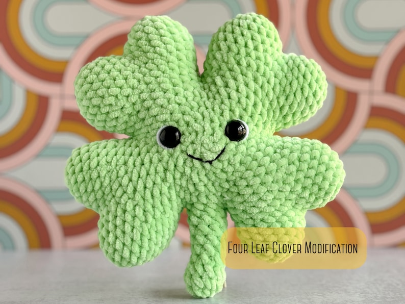 May include: A green crocheted four-leaf clover with a smiling face and black eyes. The clover is sitting on a colorful background with the text "Four Leaf Clover Modification".