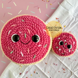 May include: Two pink crocheted donut plushies with white and green sprinkles. The larger donut has a happy face with black eyes. The smaller donut is behind the larger donut. The donuts are on a white fabric background with pink and white sprinkles.