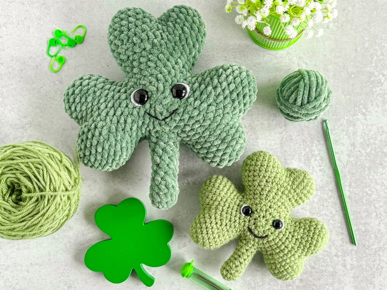 May include: Two green crocheted shamrock plushies with smiling faces and black eyes. One shamrock is larger than the other. There is green yarn, a crochet hook, and green clover-shaped embellishments in the image.