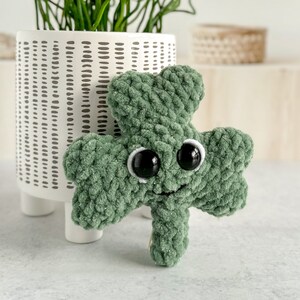 May include: A green crocheted shamrock with googly eyes and a smiling mouth. The shamrock is sitting on a white surface.