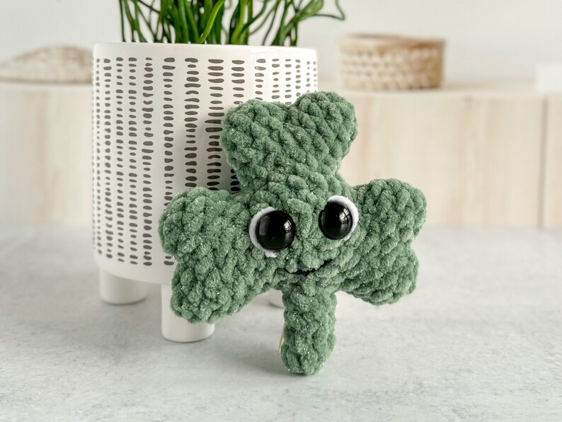May include: A green crocheted shamrock with googly eyes and a smiling mouth. The shamrock is sitting on a white surface.