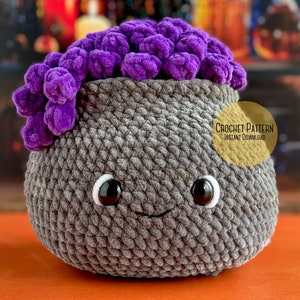 Puede incluir: Caldero de ganchillo con una parte superior de pelusa morada, ojos negros y una boca sonriente. El caldero es gris y tiene una superficie texturizada. El texto "CROCHET PATTERN INSTANT DOWNLOAD" está en un círculo amarillo en el fondo.