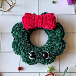 May include: A handmade green crochet wreath with a red bow on top. The wreath has two black button eyes and white stitching. The wreath is a festive decoration for the holiday season.