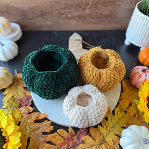 Pu&ograve; includere: Quattro ciotole fatte a mano a forma di zucca all'uncinetto nei colori verde, oro e crema. Le ciotole sono disposte su un tagliere di marmo bianco, circondate da foglie autunnali, girasoli e zucche decorative.