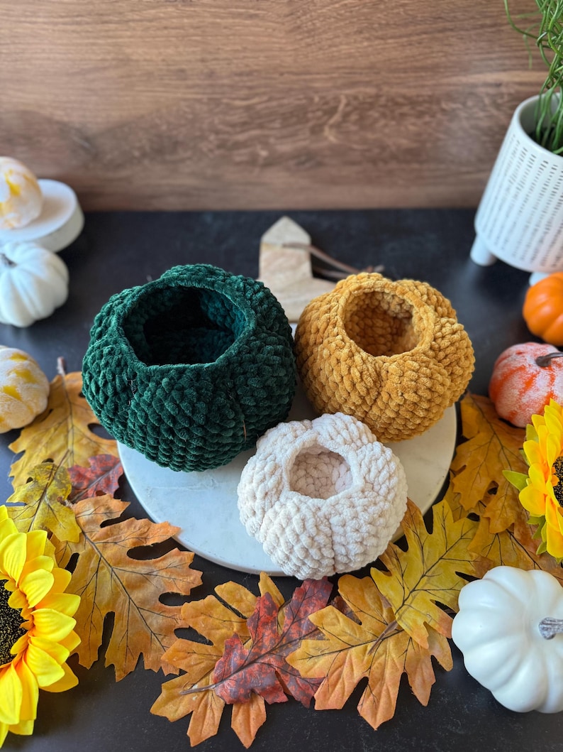 Pu&ograve; includere: Quattro ciotole fatte a mano a forma di zucca all'uncinetto nei colori verde, oro e crema. Le ciotole sono disposte su un tagliere di marmo bianco, circondate da foglie autunnali, girasoli e zucche decorative.