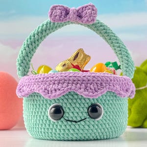 May include: A green and purple crocheted Easter basket with a bow on top. The basket is filled with chocolate eggs and candy. The basket has a smiling face with big eyes.