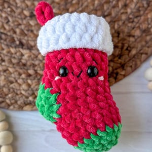 K&ouml;nnte beinhalten: Ein handgefertigter Weihnachtsstrumpf aus roter, wei&szlig;er und gr&uuml;ner Wolle. Der Strumpf hat eine wei&szlig;e Manschette und eine rote Schlaufe zum Aufh&auml;ngen. Er hat ein niedliches Gesicht mit schwarzen Augen und einem l&auml;chelnden Mund. Der Boden ist gr&uuml;n.