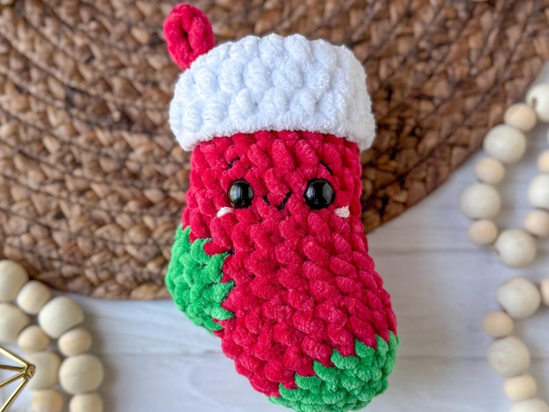 K&ouml;nnte beinhalten: Ein handgefertigter Weihnachtsstrumpf aus roter, wei&szlig;er und gr&uuml;ner Wolle. Der Strumpf hat eine wei&szlig;e Manschette und eine rote Schlaufe zum Aufh&auml;ngen. Er hat ein niedliches Gesicht mit schwarzen Augen und einem l&auml;chelnden Mund. Der Boden ist gr&uuml;n.