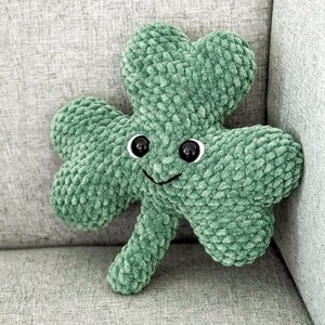 May include: A green plush shamrock with a smiling face and black eyes. The shamrock is sitting on a gray couch.