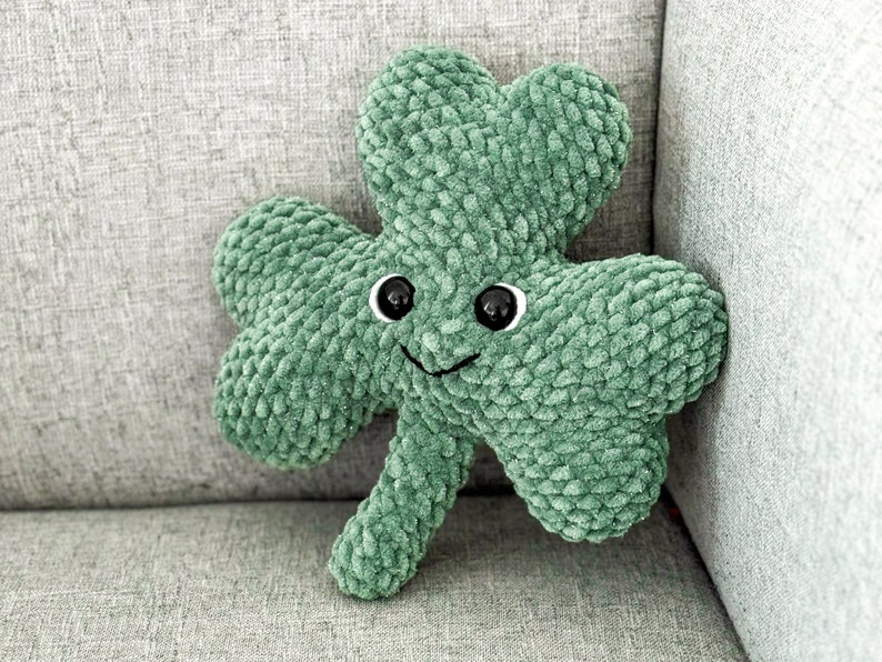 May include: A green plush shamrock with a smiling face and black eyes. The shamrock is sitting on a gray couch.