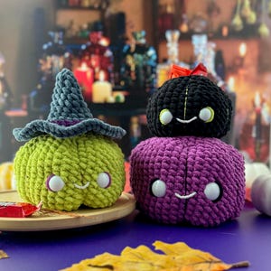Pu&ograve; includere: Tre zucche di Halloween all'uncinetto. Una &egrave; verde con un cappello da strega, un'altra &egrave; nera con un fiocco rosso e la terza &egrave; viola. Ognuna ha una faccia sorridente con occhi bianchi. Sono su un vassoio di legno.