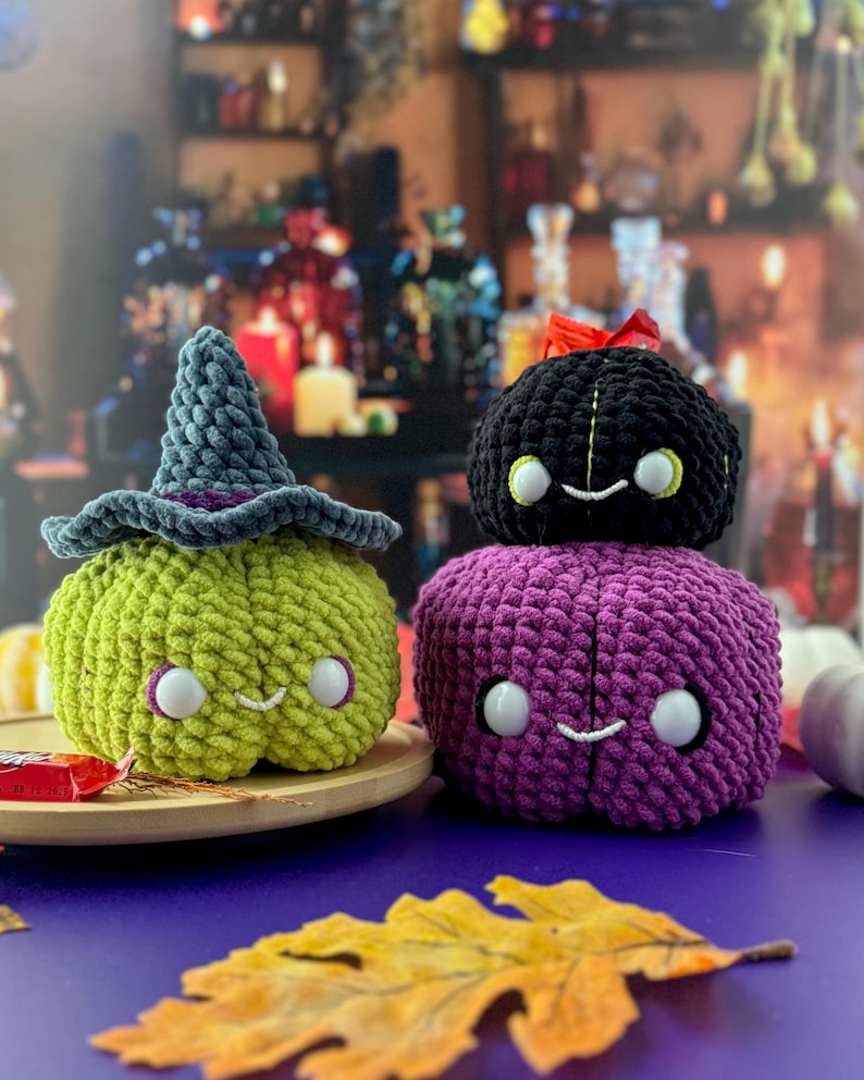 Pu&ograve; includere: Tre zucche di Halloween all'uncinetto. Una &egrave; verde con un cappello da strega, un'altra &egrave; nera con un fiocco rosso e la terza &egrave; viola. Ognuna ha una faccia sorridente con occhi bianchi. Sono su un vassoio di legno.