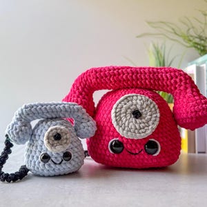 May include: Two crocheted toy telephones. One is light blue, the other is bright pink. Both have a circular gray and black detail, black eyes, and a smiling mouth. The pink phone is larger. The light blue phone has a black cord.