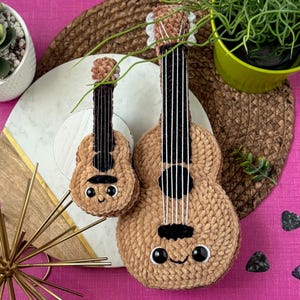 Könnte beinhalten: Zwei Plüsch-Ukulele-Spielzeuge in Beige mit schwarzen Saiten und lächelnden Gesichtern. Eines ist größer als das andere. Die Ukulelen befinden sich auf einem weißen und goldenen Dekoteller, mit einem rosa Hintergrund und einer kleinen Sukkulentenpflanze.