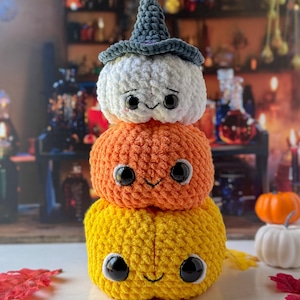 Pu&ograve; includere: Una pila di tre zucche all'uncinetto nei toni del giallo, arancione e bianco, sormontate da un cappello da strega grigio. Ogni zucca ha grandi occhi neri e un sorriso cucito. Una decorazione festosa per Halloween.