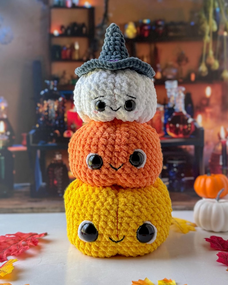 Pu&ograve; includere: Una pila di tre zucche all'uncinetto nei toni del giallo, arancione e bianco, sormontate da un cappello da strega grigio. Ogni zucca ha grandi occhi neri e un sorriso cucito. Una decorazione festosa per Halloween.