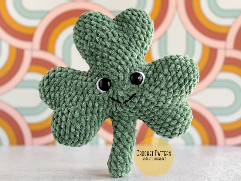 May include: Green crocheted shamrock with a smiling face and black eyes. The shamrock is sitting on a white surface with a colorful background. Crochet Pattern Instant Download.