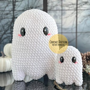 Könnte beinhalten: Zwei gehäkelte Geister-Amigurumi-Spielzeuge, eines groß und eines klein, mit schwarzen Augen und rosa Wangen. Der größere Geist ist weiß und der kleinere Geist ist weiß. Die Geister sitzen auf einer dunklen Oberfläche mit einer Kiefernzapfen und einem verschwommenen Hintergrund aus Bäumen.
