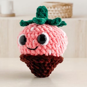 Peut inclure: Un jouet en peluche en forme de fraise crochet&eacute; avec un dessus vert feuillu, un fond brun chocolat et de grands yeux noirs.