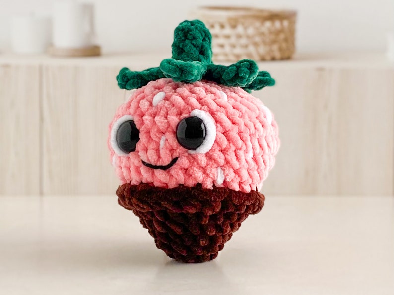 Peut inclure: Un jouet en peluche en forme de fraise crochet&eacute; avec un dessus vert feuillu, un fond brun chocolat et de grands yeux noirs.