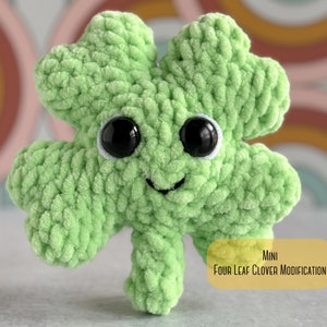 May include: A small green crocheted four-leaf clover with a smiling face and black eyes. The clover is sitting on a white surface with a rainbow background. MINI FOUR LEAF CLOVER MODIFICATION