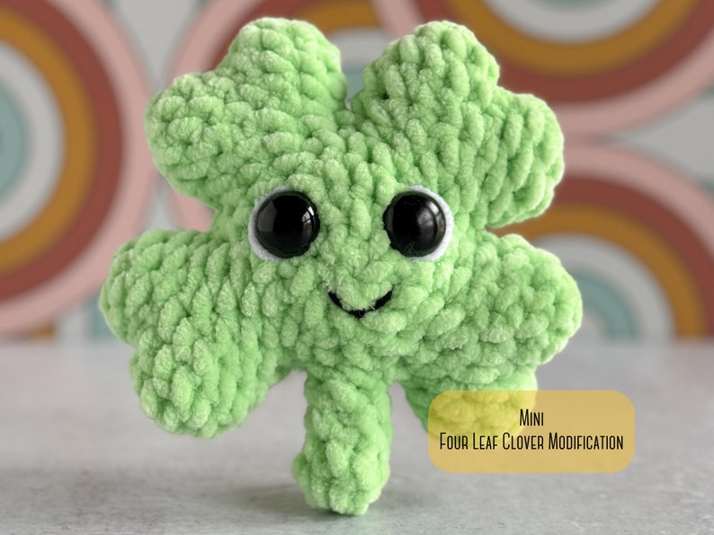 May include: A small green crocheted four-leaf clover with a smiling face and black eyes. The clover is sitting on a white surface with a rainbow background. MINI FOUR LEAF CLOVER MODIFICATION
