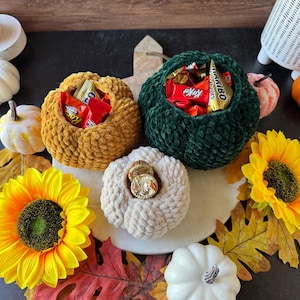 Pu&ograve; includere: Una composizione a tema autunnale con tre ciotole a forma di zucca all'uncinetto piene di caramelle. Le zucche sono nei toni dell'oro, del verde e del crema, circondate da girasoli, foglie decorative e piccole zucche.