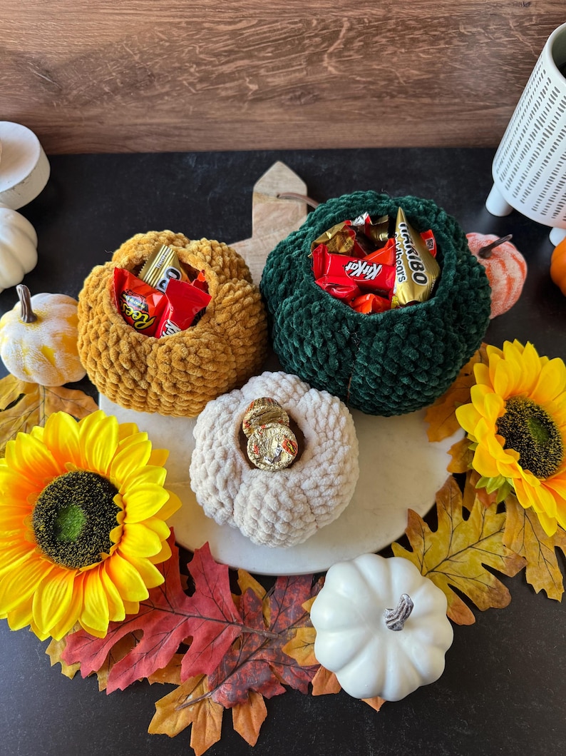Pu&ograve; includere: Una composizione a tema autunnale con tre ciotole a forma di zucca all'uncinetto piene di caramelle. Le zucche sono nei toni dell'oro, del verde e del crema, circondate da girasoli, foglie decorative e piccole zucche.