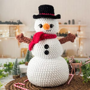 Op de afbeelding: Een handgemaakte sneeuwpop met een zwarte hoge hoed, een rode sjaal en bruine takarmen. De sneeuwpop heeft een wortelneus, zwarte knoopogen en een lachende mond. Hij is gemaakt van wit garen en staat op een bruine geweven mat.