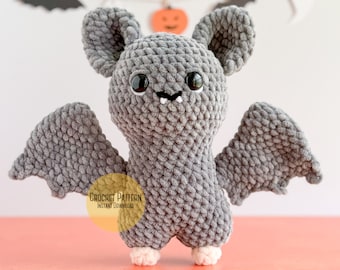 May include: Grey crocheted bat amigurumi toy with black eyes and white teeth. The bat has large grey wings and is standing on a pink surface. Crochet pattern instant download.