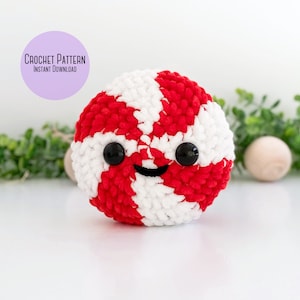 May include: A red and white crocheted candy cane with a smiling face. The candy cane is sitting on a white surface with green foliage in the background. Crochet Pattern Instant Download.