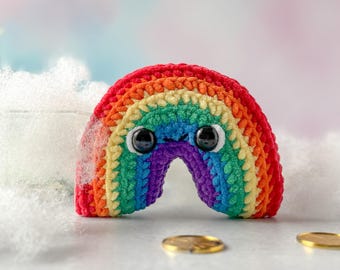 Puede incluir: Un arcoíris tejido a ganchillo con una cara sonriente y grandes ojos negros. El arcoíris presenta colores vibrantes, incluyendo rojo, naranja, amarillo, verde, azul y morado. El artículo artesanal está sobre un fondo suave y borroso con monedas de oro.
