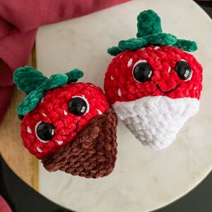 Peut inclure: Deux jouets en peluche en forme de fraise au crochet. Une fraise est rouge avec un fond en chocolat blanc et l'autre est rouge avec un fond en chocolat brun. Les deux fraises ont des feuilles vertes et des yeux noirs.