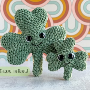May include: Two green crocheted shamrock plushies with black eyes and smiling faces. The larger shamrock is standing behind the smaller shamrock. The background is a colorful pattern of pink, orange, and blue.
