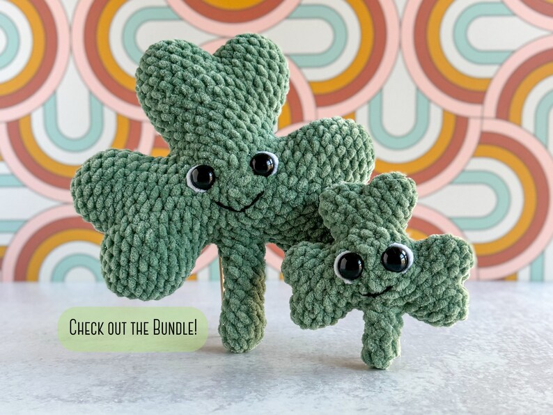 May include: Two green crocheted shamrock plushies with black eyes and smiling faces. The larger shamrock is standing behind the smaller shamrock. The background is a colorful pattern of pink, orange, and blue.