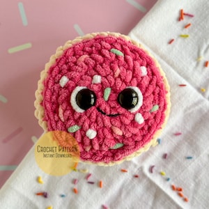 Może przedstawiać: Różowy, szydełkowy ciasteczko z białymi i zielonymi posypkami i czarnymi oczami. Ciasteczko leży na białym tle z kolorowymi posypkami.