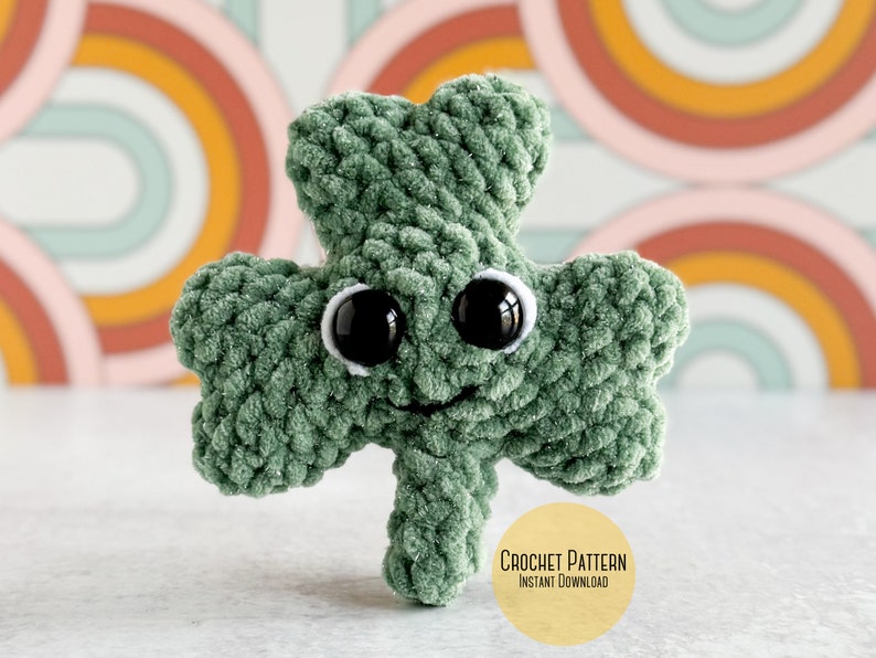 May include: A green crocheted shamrock with googly eyes and a smiling mouth. The shamrock is sitting on a white surface with a colorful background of rainbow arches. The text "CROCHET PATTERN INSTANT DOWNLOAD" is on a yellow circle.