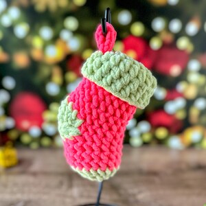 K&ouml;nnte beinhalten: Ein kleiner, handgefertigter Weihnachtsstrumpf in leuchtendem Rot mit gr&uuml;ner Manschette und gr&uuml;nem Bl&auml;tterakzent. Der Strumpf ist aus einem groben, strukturierten Garn gefertigt und h&auml;ngt an einem schwarzen Metallst&auml;nder. Der Hintergrund ist mit Weihnachtsbeleuchtung verschwommen.