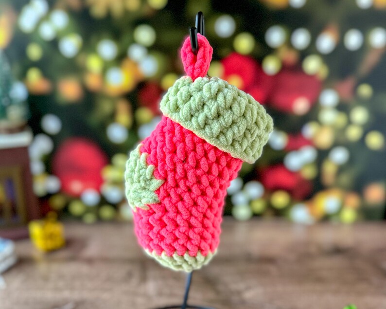 K&ouml;nnte beinhalten: Ein kleiner, handgefertigter Weihnachtsstrumpf in leuchtendem Rot mit gr&uuml;ner Manschette und gr&uuml;nem Bl&auml;tterakzent. Der Strumpf ist aus einem groben, strukturierten Garn gefertigt und h&auml;ngt an einem schwarzen Metallst&auml;nder. Der Hintergrund ist mit Weihnachtsbeleuchtung verschwommen.