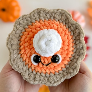 May include: A crocheted pumpkin pie plush toy with a white whipped cream topping and googly eyes. The pie is orange and brown and has a cute, smiling face.