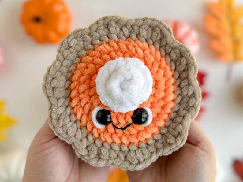May include: A crocheted pumpkin pie plush toy with a white whipped cream topping and googly eyes. The pie is orange and brown and has a cute, smiling face.